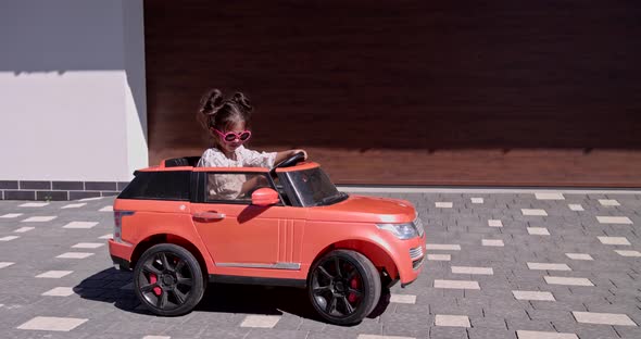 Little Girl Sits In A Toy Car Near The Garage