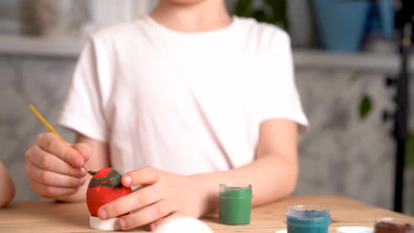 Baby Decorates The Easter Egg with Paints and Brush Sitting at the Table Hands Closeup