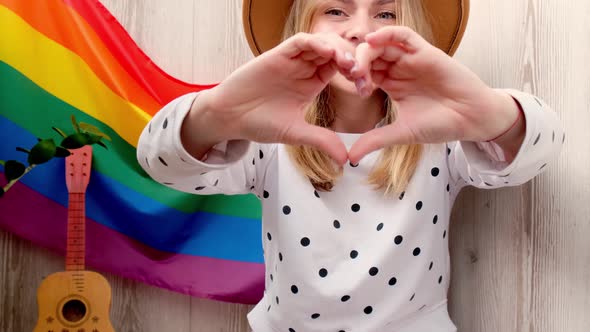 Young Blonde Millennial Hippie Woman Showing Heart Sign with Hands with Rainbow LGBTQ Flag at Home