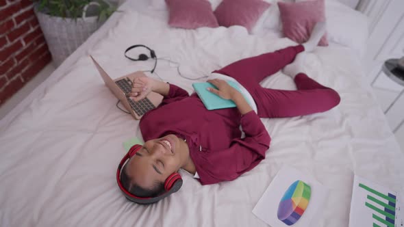 High Angle View of Joyful Young African American Woman with Toothy Smile Lying on Bed Enjoying Music