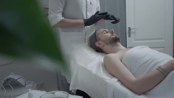 Relaxed Man Lying on Beauty Bed As Unrecognizable Beautician in Gloves Applying Moisturizing Facial