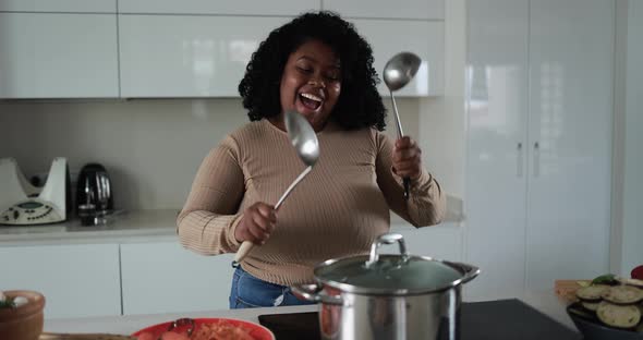 Black young african woman having fun cooking and dancing inside kitchen at home