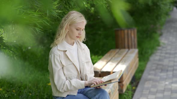 A Cute Young Woman Sits on a Park Bench and Reads a Book Carefree