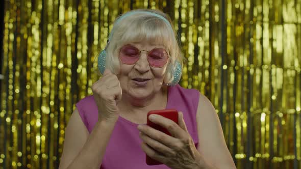 Stylish Happy Senior Old Woman Holding Cell Phone, Listening To Music in Headphones and Dancing