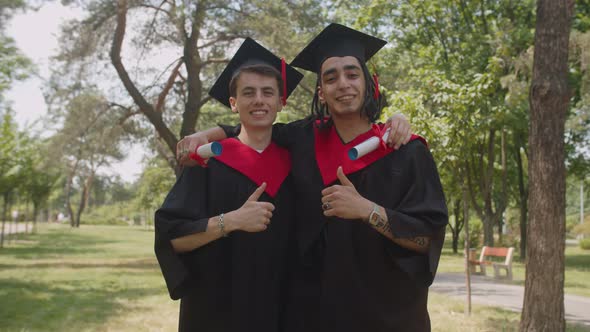 Smiling Multiethnic Male Graduates with Diplomas Showing Thumbs Up Outdoors