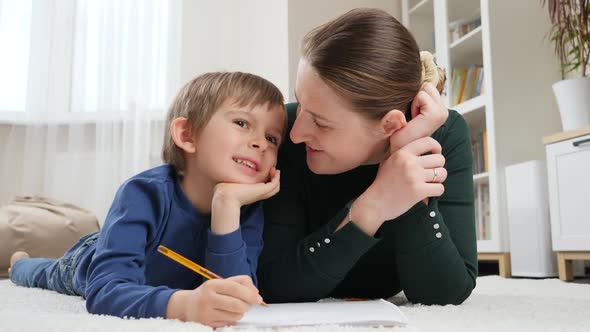 Little Boy and Young Mother Lying on Floor in Living Room and Doing Homework in Copybook