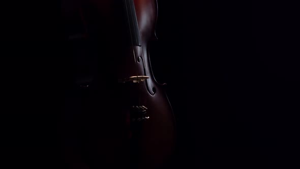 Cello, dimly lit, makes half turn on dark background in studio, front view. Cello is spinning