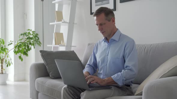 Smiling Middle Aged Man Using Laptop Computer Sitting on Couch at Home