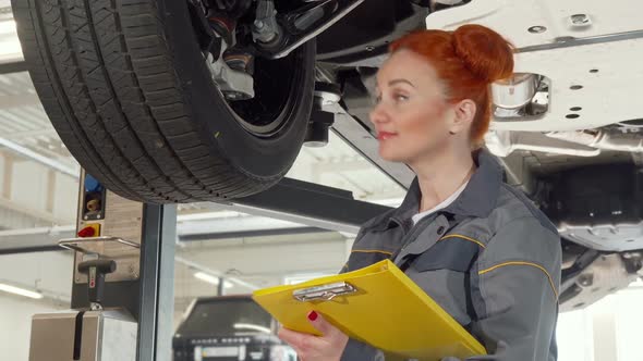 Female Car Mechanic Examining Wheels of a Car on a Lift, Taking Notes on Clipboard