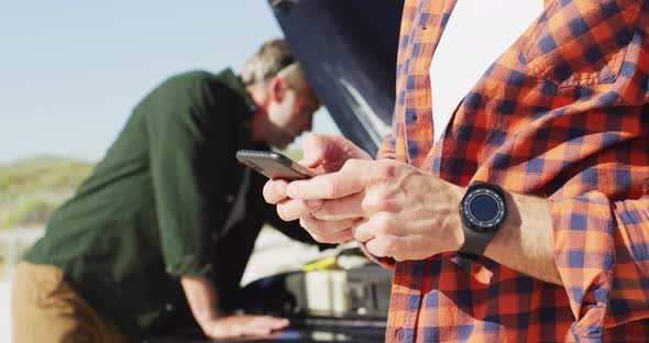 Stressed caucasian gay male couple standing by broken down car using smartphone at the beach