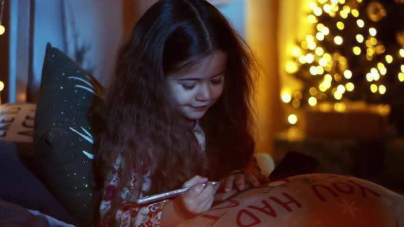 Cute Little Girl While Lying on Bed Write Letter To Santa in Room Decorated for Christmas, Makes a