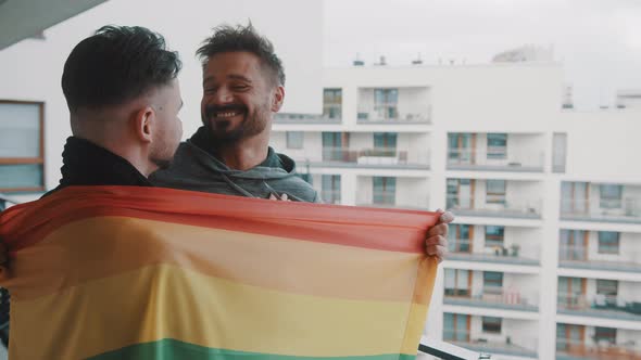 Young Excited Gay Male Couple Standing on the Balcony Covered with Rainbow Flag