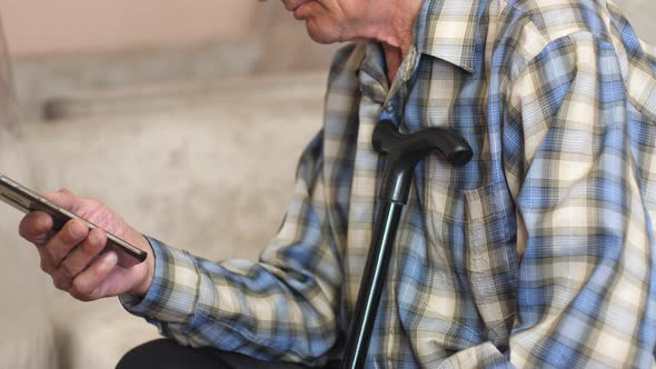 Focused serious caucasian pensioner sits with a walking stick and uses a smartphone. An elderly man 