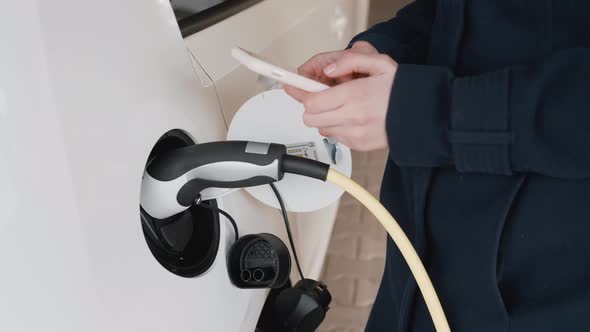 Woman Standing Near the Charging Electric Car and Scrolling Her Smartphone