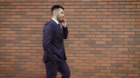 Businessman Dressed in Shirt Talking Smartphone While Walks on the Background of a Modern Brick Wall