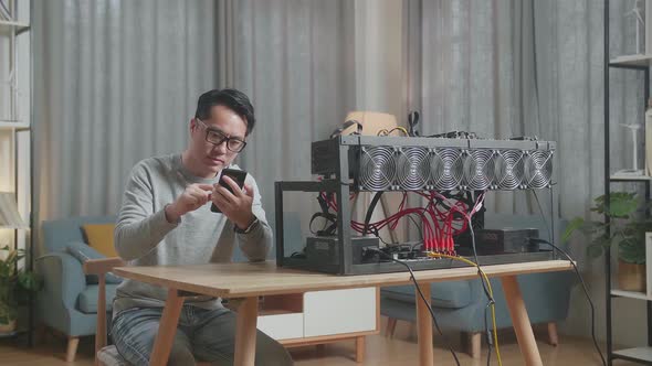 Asian Man Using Smartphone And Looking At The Mining Rig For Mining Cryptocurrency