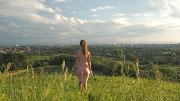 Young woman in pink dress walking on green summer field outdoors.