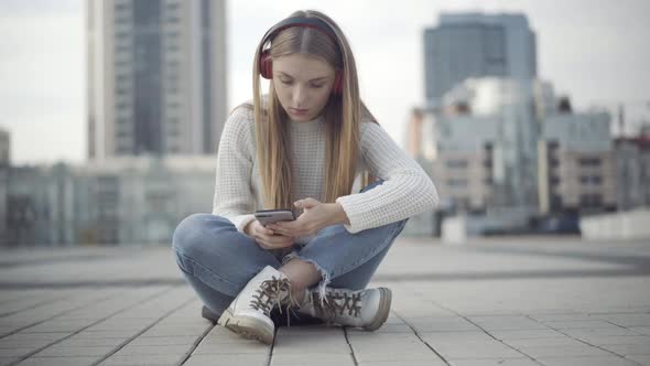 Young Relaxed Woman Sitting on City Square Listening To Music in Headphones and Scrolling Smartphone