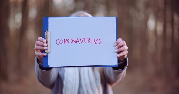 Woman with Protective Mask Holding Coronavirus Inscription in Hands in Forest