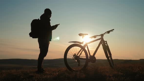 Silhouette Cyclist Use Mobile Phone on a Mountain at Sunset. alt
