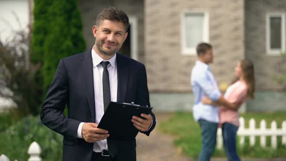 Smiling Broker Standing With Documents, Happy Family Hugging Near Their New Home