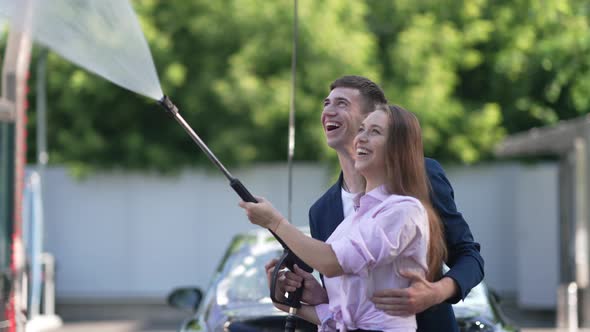 Young Couple Laughing Turning on High Pressure Washer Standing at Car Wash Service Outdoors on Sunny