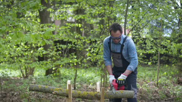 Handsome Lumberjack in Working Uniform Sawing Wood with Chainsaw on Sawhorse