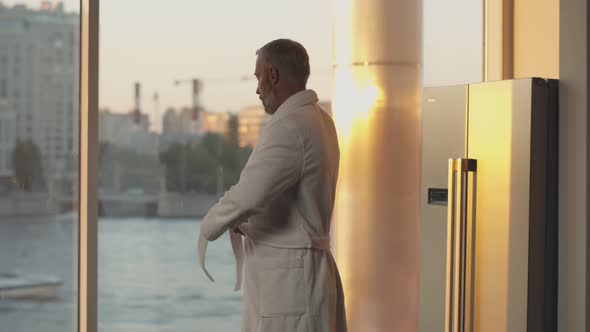 Senior Man in Bathrobe Looking Out of Window Enjoying Sunset Cityscape at Home