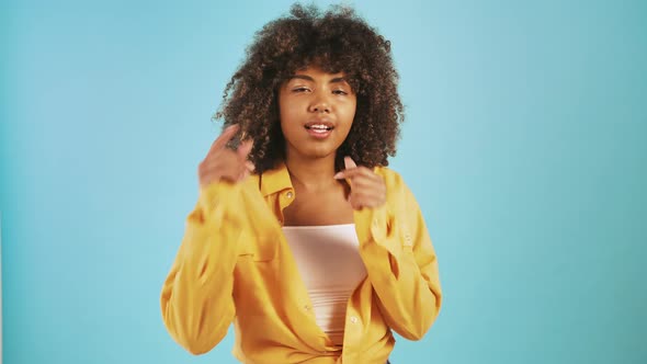 Young Afro American Female is Smiling and Pointing at you By Forefingers While Posing Against Blue