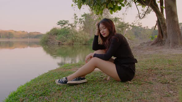 Thai Young Woman Sits Pensively By Lake in Park at Sunset