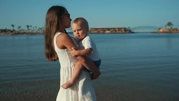 Long Hair Teenage Girl Walking Along Sunny Seaside with Baby Boy on Hands.