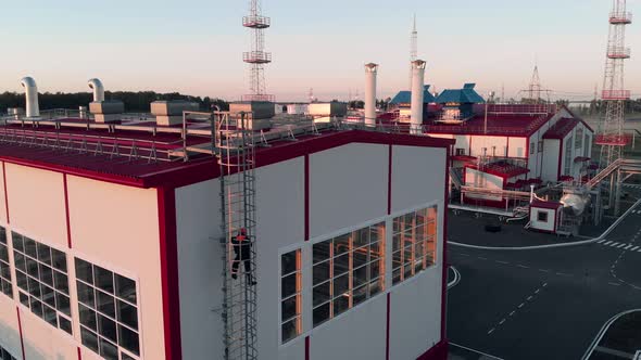 A Drone Flies Up to a Worker in Overalls and a Helmet Who Climbs the Stairs to an Industrial