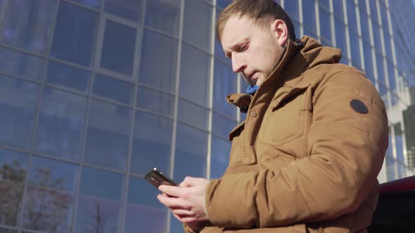 Young Man Sitting By the Car Using His Smart Phone Modern Building with Big Windows at the