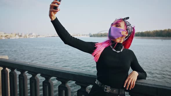 Hipster Female in Informal Clothes and Protective Mask is Taking Selfie on Smartphone Leaning on