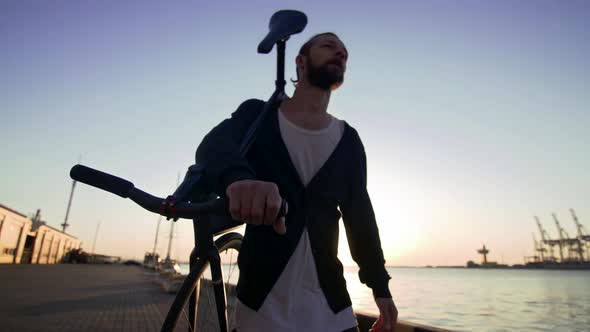 Stylish Handsome Man Walking with Bicycle on His Shoulder During Sunset or Sunrise with Sea Port on