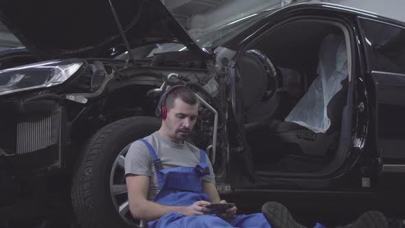 Portrait of Young Caucasian Male Auto Mechanic in Headphones Leaning Against Car Wheel and Using