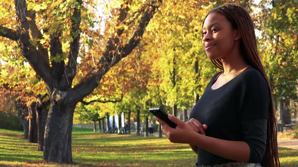 Young Beautiful Black Woman Works on Smartphone in the Autumn Park