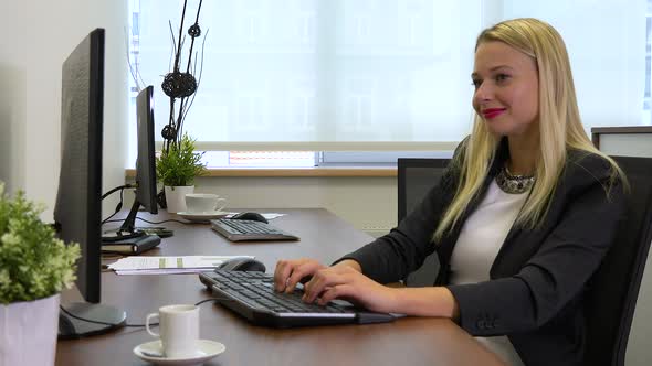 An office worker sits at a desk and works on a computer, eventually smiles at the camera