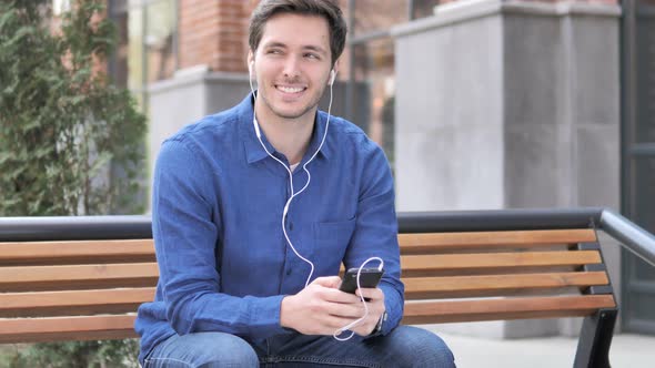 Young Man Listening Music on Smartphone while Sitting on Bench