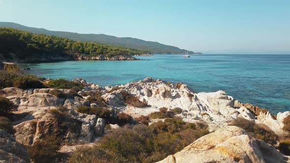 Aegean sea coast with greenery around, rocks, bushes and trees, blue water, moving yacht, Greece. Sl