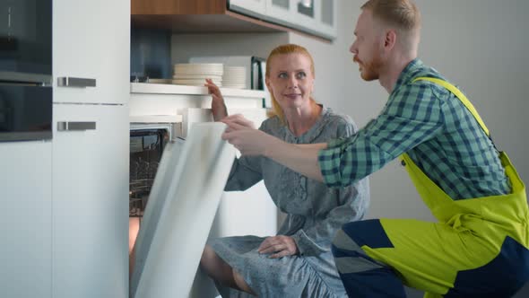 Young Service Worker Repairing Dishwasher Appliance In Kitchen Shaking Hands with Mature Housewife