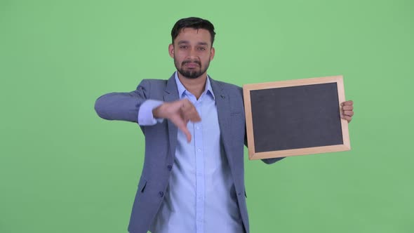 Stressed Young Bearded Persian Businessman Holding Blackboard and Giving Thumbs Down