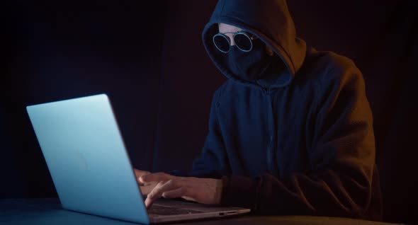 A hacker with a closed face sits at a laptop in a dark room