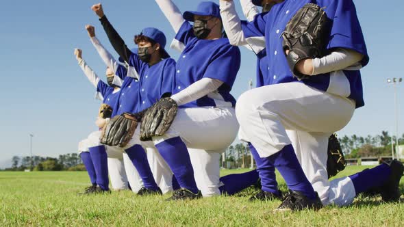 Diverse team of female baseball players in face masks kneeling in line with arms raised