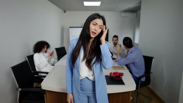 Exhausted Asian Slim Woman Rubbing Temples and Yawning Standing in Office with Blurred Coworkers at