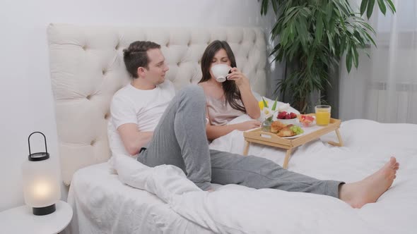 Happy Young Couple Eating Breakfast in Bed and Drinking a Cup of Coffee