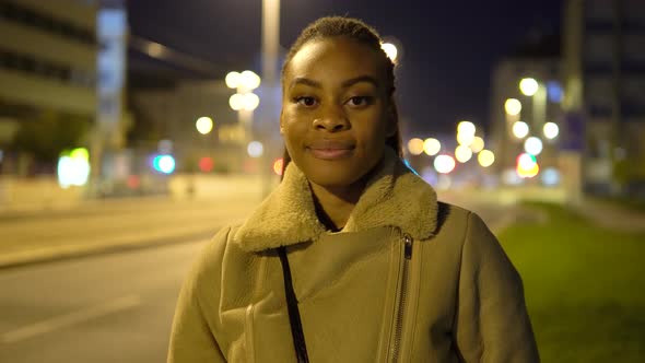 A Young Black Woman Smiles at the Camera in a Street in an Urban Area at Night