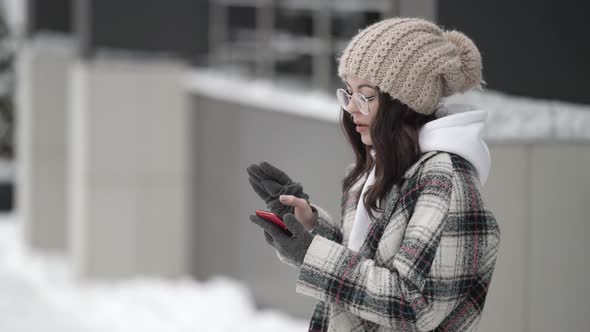 A Young Darkhaired Woman in Round Glasses and a Knitted Hat Stands Outside on a Winter Day Looking