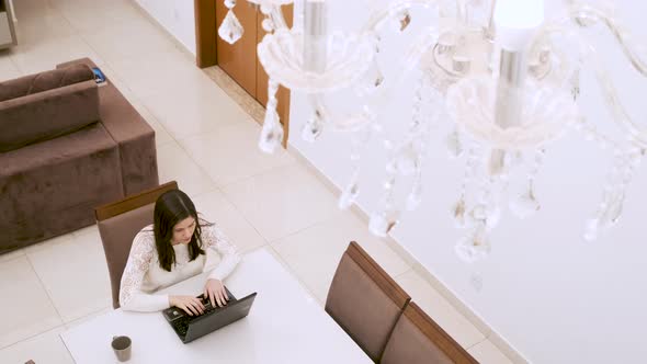 Caucasian woman drinks coffee and types on laptop at homework, under chandelier. Shot down. Homework