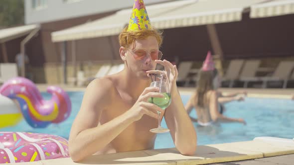 Positive Redhead Caucasian Man in Party Hat Drinking Cocktail in Pool and Showing Thumb Up. Portrait